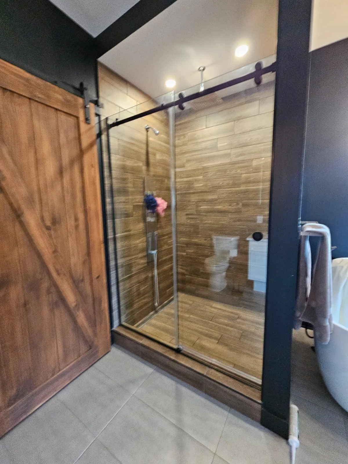 A modern walk-in shower with wooden wall tiles, a glass door, and a handheld showerhead, in a bathroom with gray tiled flooring.