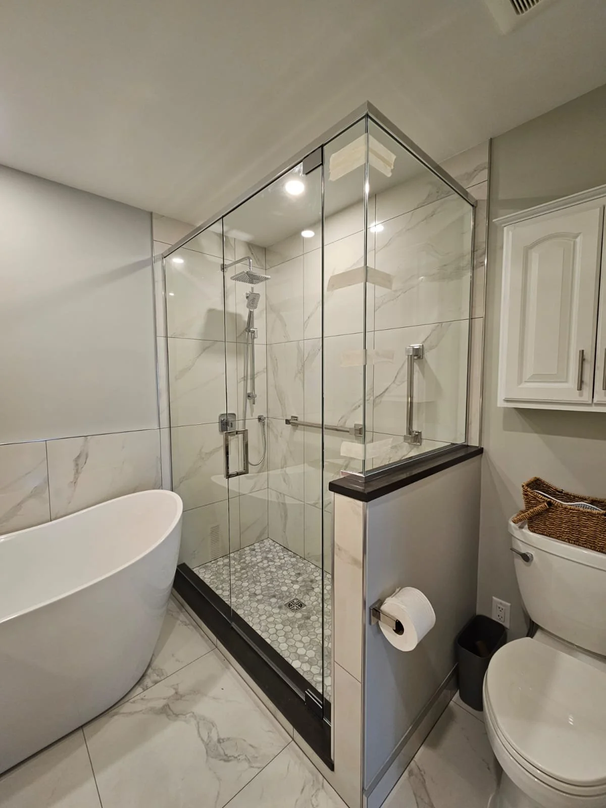 A modern bathroom with a corner glass-shower, a white freestanding bathtub, a toilet with a wicker basket on top, and white cabinets.