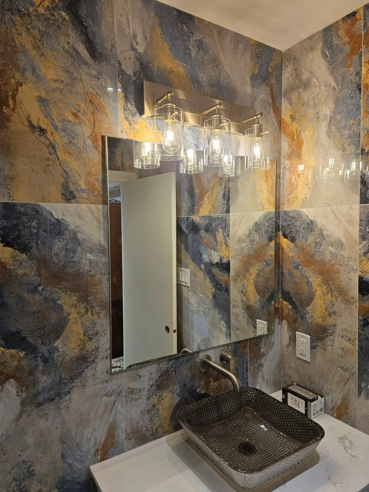 Bathroom sink with a black square vessel basin, wall-mounted faucet, marble countertop, decorative wall tiles, a large mirror, a light fixture with multiple glass shades, and some electrical outlets.
