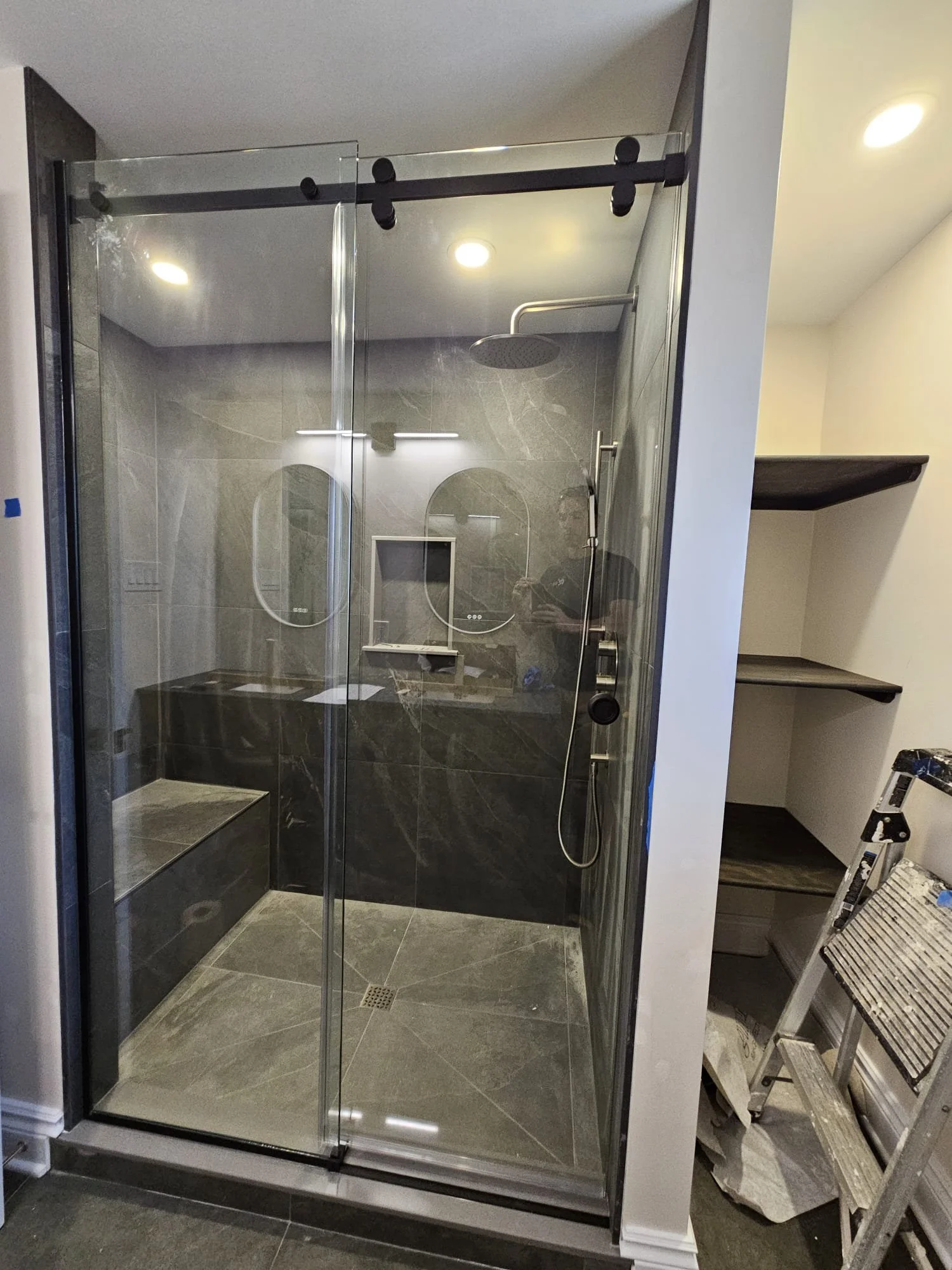 Modern glass shower stall with black tile walls and a bench, featuring a ceiling showerhead and a handheld shower, next to wall-mounted shelves and construction tools.
