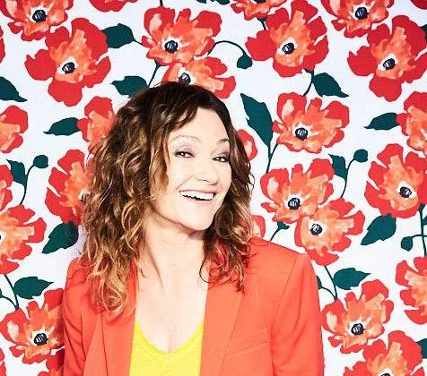 Woman with curly brown hair, wearing an orange blazer and yellow top, smiling in front of a floral wallpaper featuring red poppies with dark centers and dark green leaves.