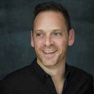 A smiling man with short brown hair, wearing a black shirt, looking to the side with a dark background.