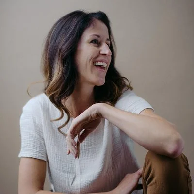A woman with dark wavy hair smiling and looking to the side, wearing a white t-shirt and sitting against a neutral background.