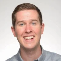 A smiling man with short brown hair, wearing a blue shirt, against a plain white background.