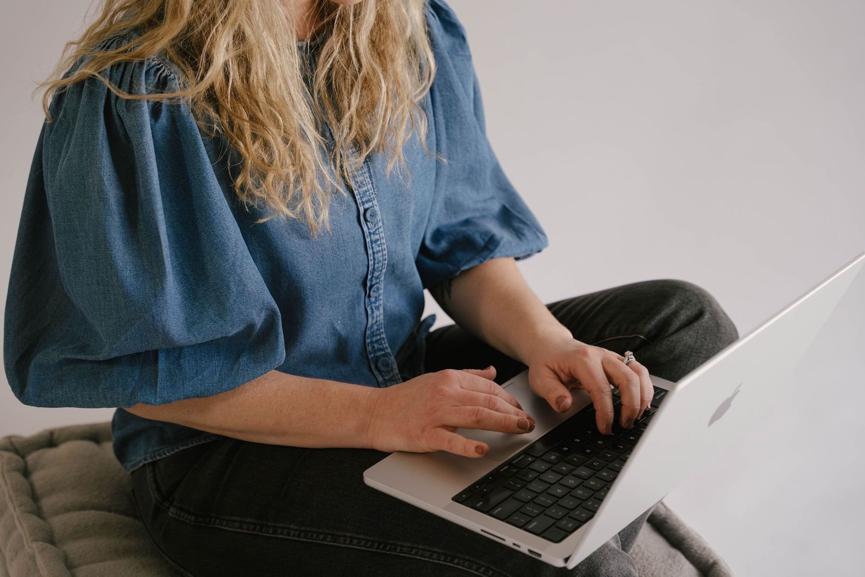 A woman sitting cross-legged on a cushioned surface, using a silver MacBook laptop. She is wearing a denim shirt and dark jeans.