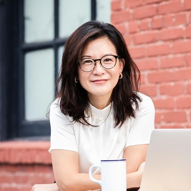 A woman with shoulder-length dark hair, glasses, and hoop earrings, smiling at the camera, sitting outdoors in front of a brick wall, with a laptop and a white mug on the table.