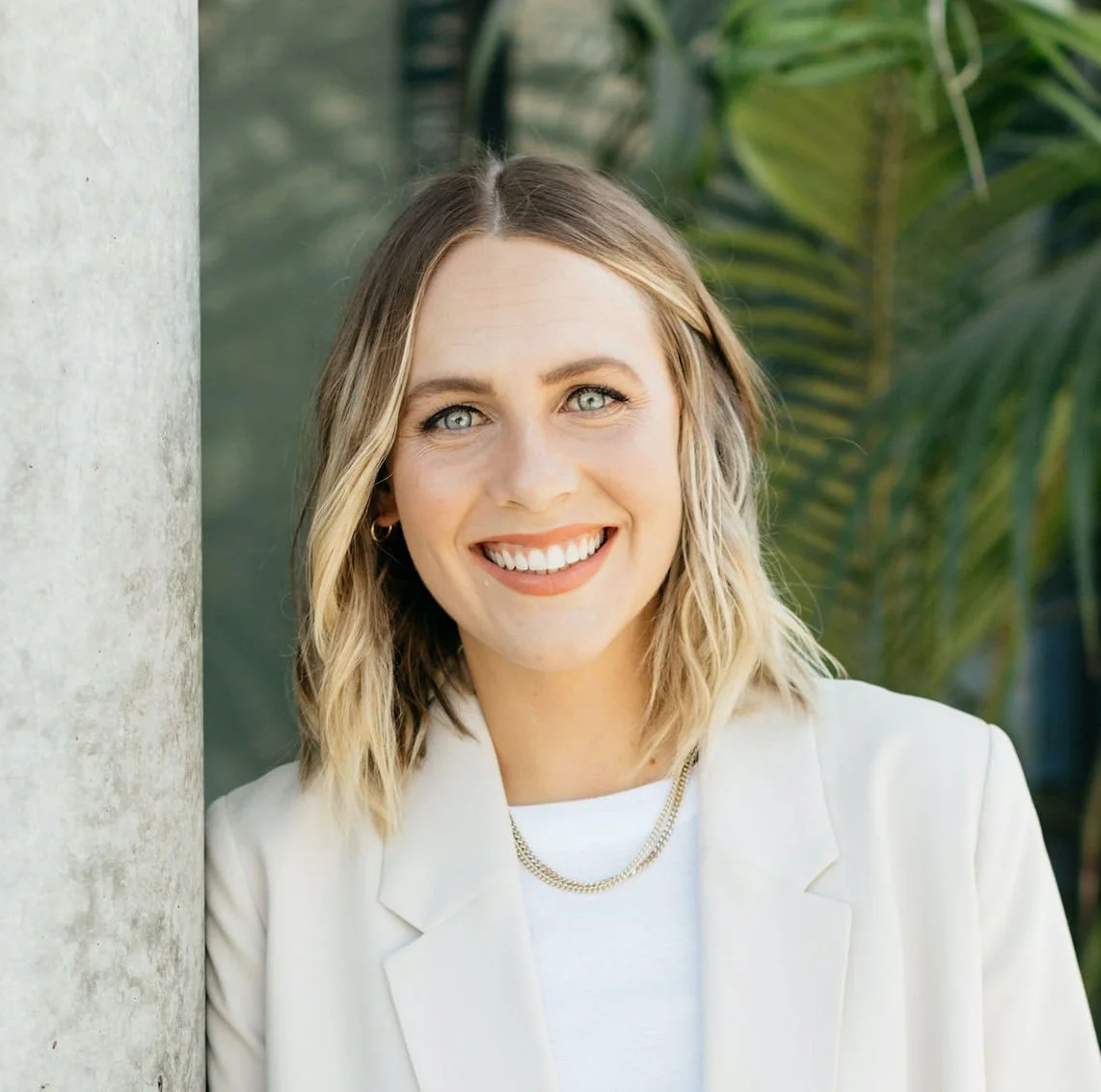 A smiling woman with shoulder-length blonde hair, blue eyes, wearing a white blazer and gold jewelry, standing outdoors with greenery in the background.
