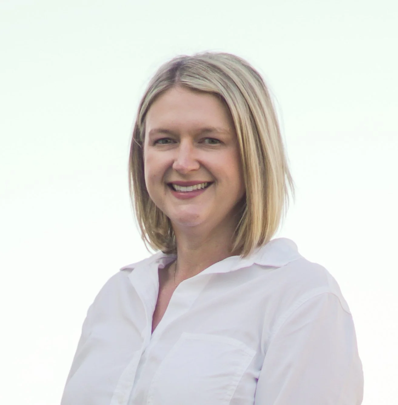 Portrait of a woman with shoulder-length blonde hair, smiling, wearing a white collared shirt, against a bright outdoor background.