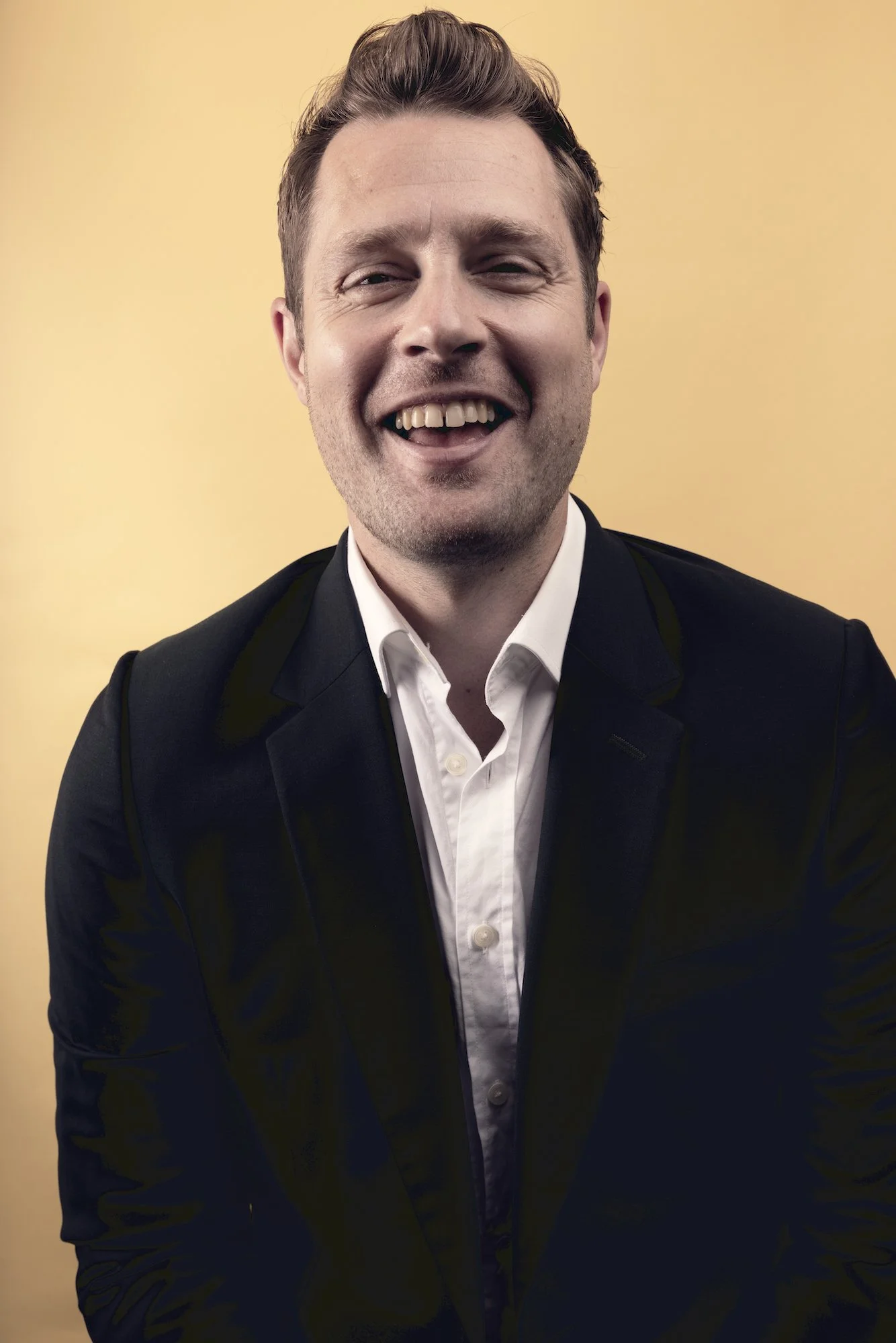 A man with light skin, short brown hair, wearing a dark blazer over a white shirt, smiling and looking happy, standing against a plain yellow background.