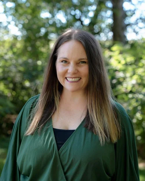 A woman with long brown hair smiling outdoors, wearing a dark green graduation gown in front of a background of trees and green foliage.