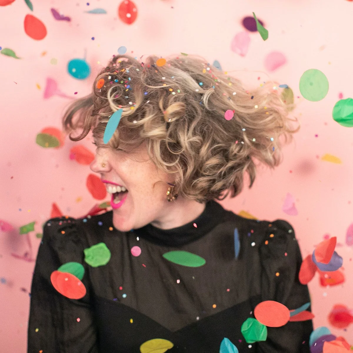 A woman with curly blonde hair is laughing and turning her head, surrounded by colorful confetti falling around her. She is wearing a black top with sheer sections and pink lipstick, against a pink background.