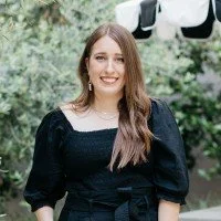 Portrait of a smiling woman with long brown hair, wearing a black outfit, outdoors with green foliage in the background.