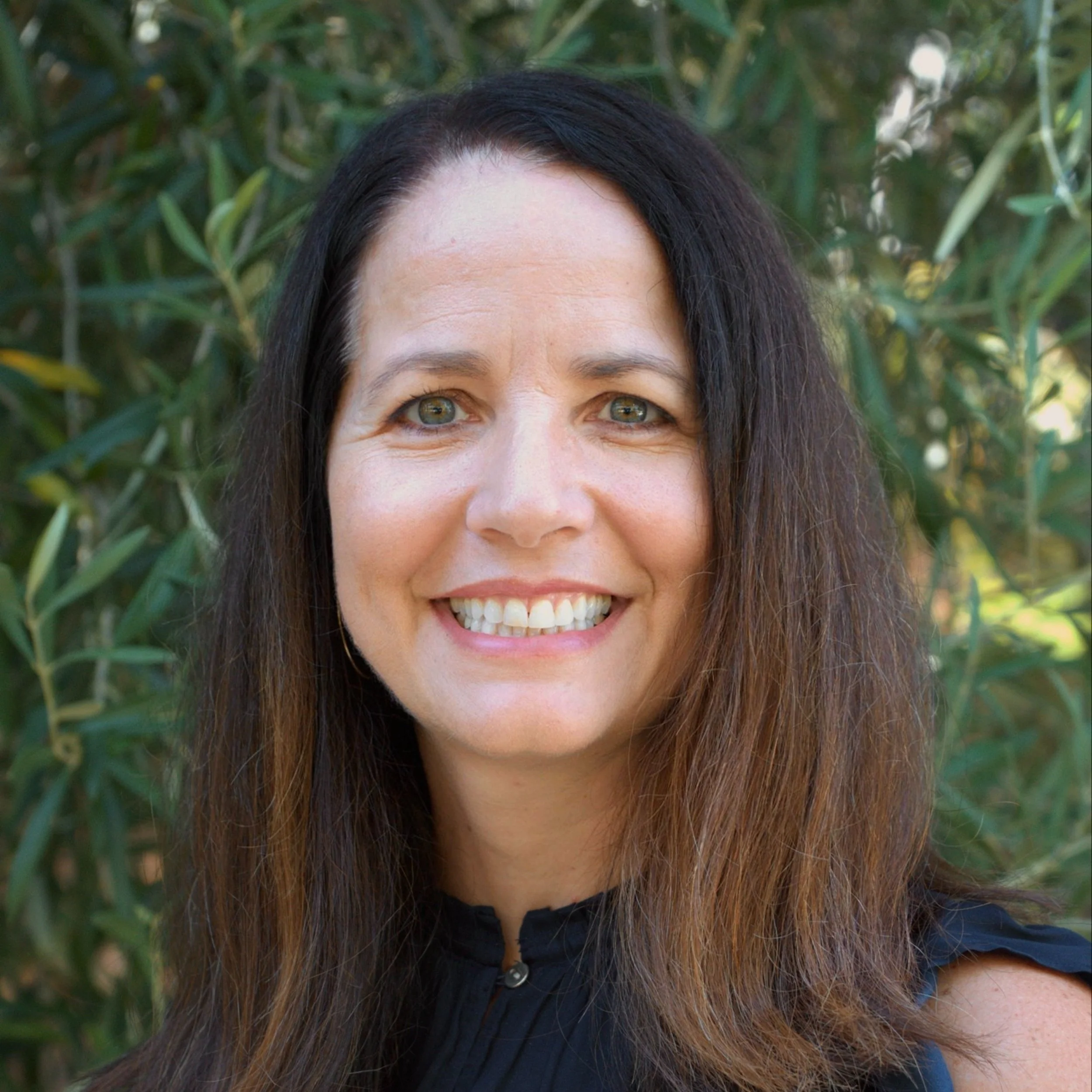 Smiling woman with long dark hair, wearing a black top, standing outdoors in front of green foliage.