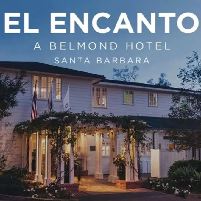 The entrance of El Encanto hotel in Santa Barbara at dusk, showing a white building with lit windows, American flags, and lush greenery.