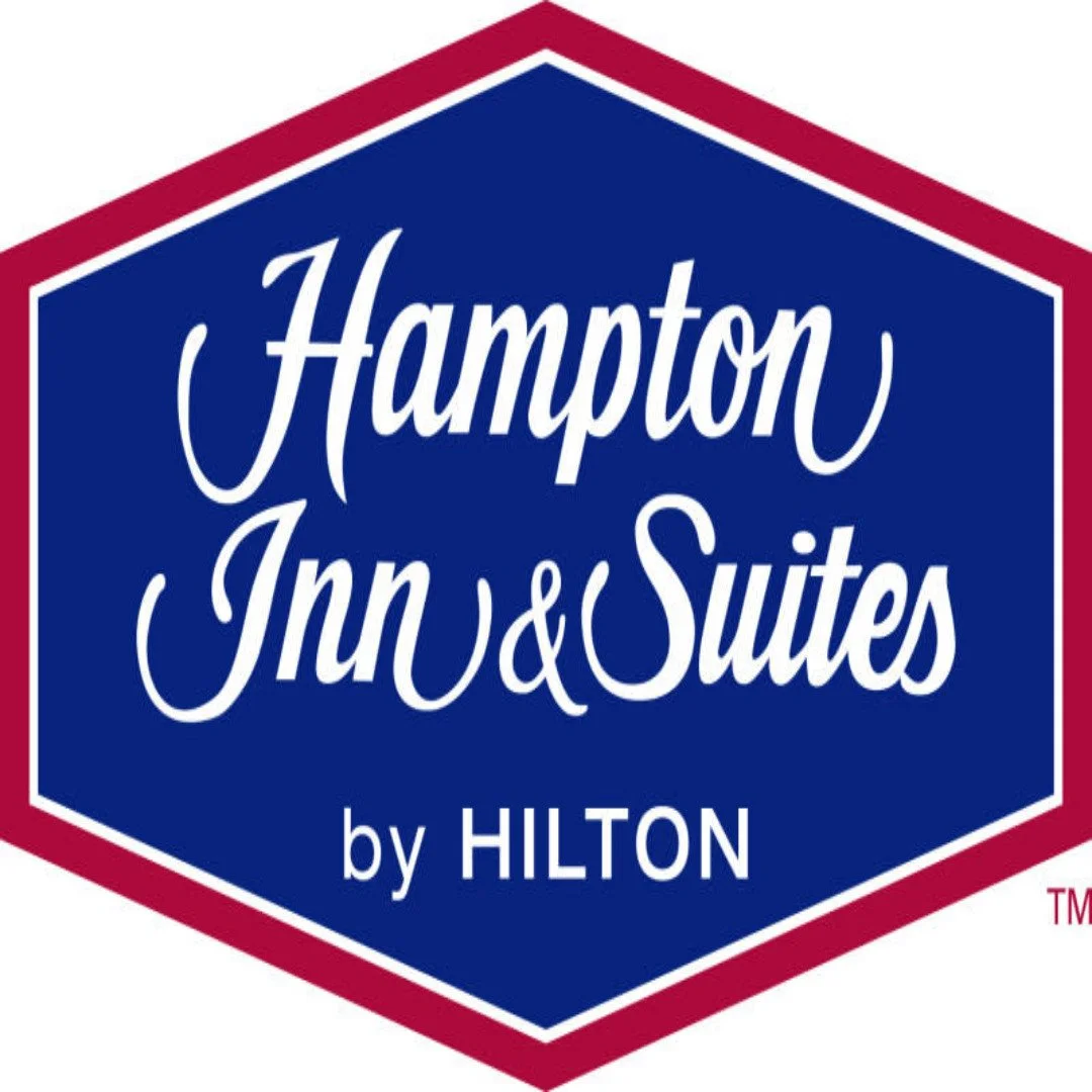 Logo of Hampton Inn & Suites by Hilton with a blue shield shape, red border, and white text.