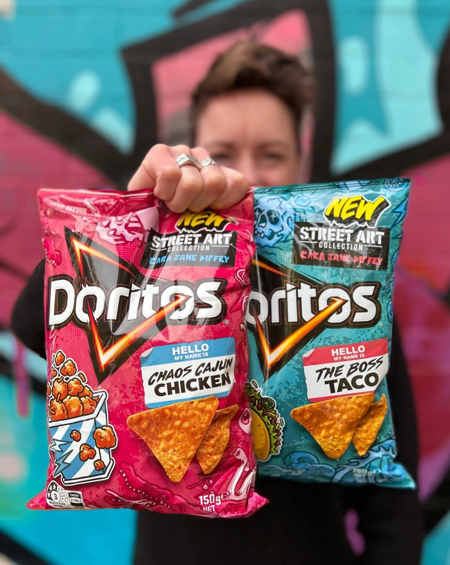 Person holding two bags of Doritos chips, one pink with a label 'Chaos Cajun Chicken' and the other blue with a label 'The Boss Taco', in front of a colorful graffiti wall