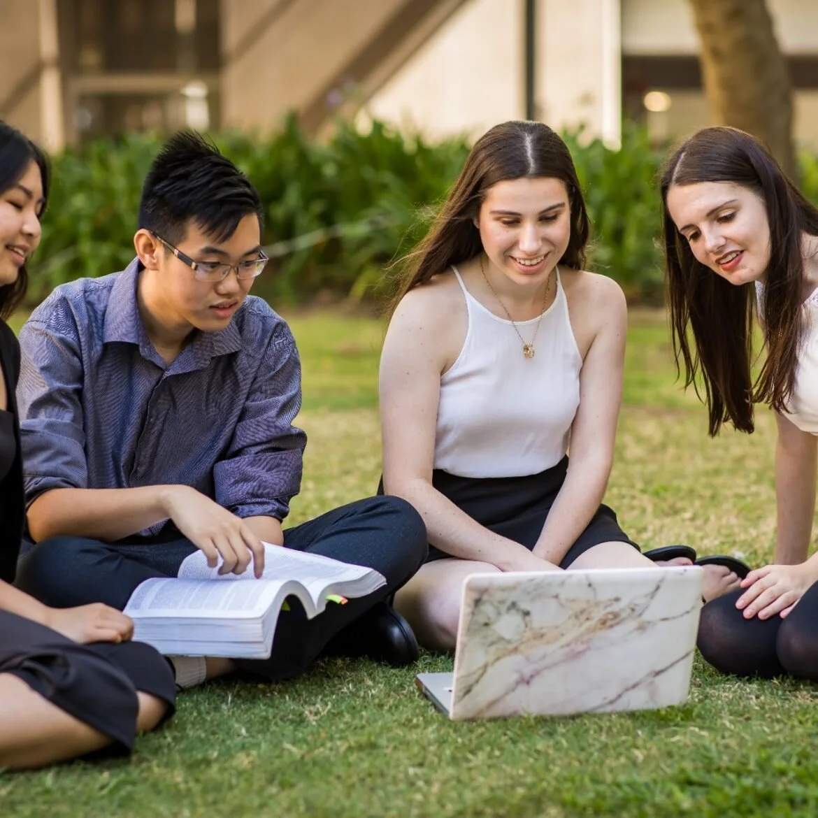 Group of four young adults sitting on grass outdoors, looking at a laptop and reading a book together, during daytime.