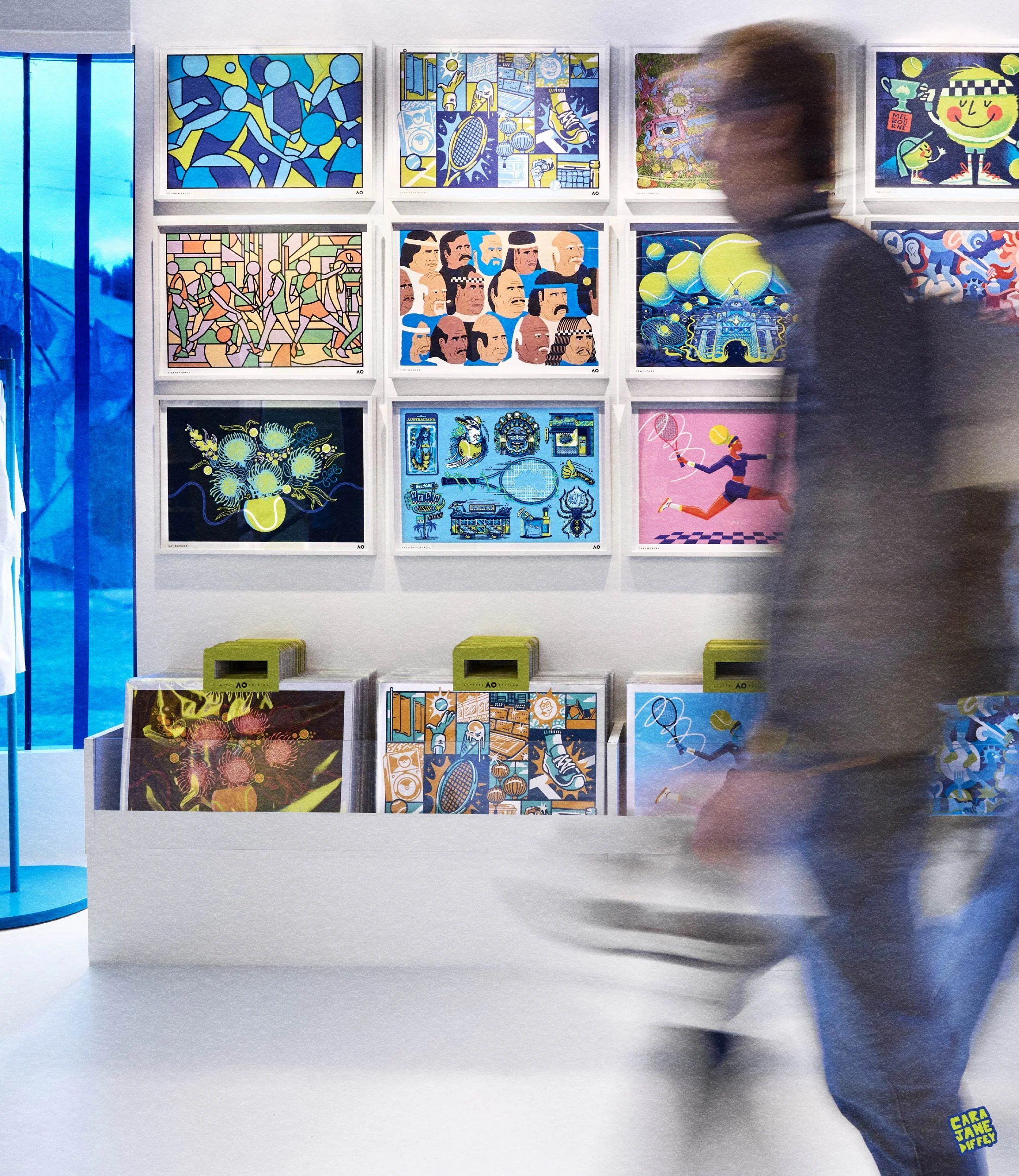 An art gallery display consists of colorful abstract and illustrative artworks on a white wall, with a person walking past, blurred due to motion, and a white table holding more framed pieces in front of the wall.