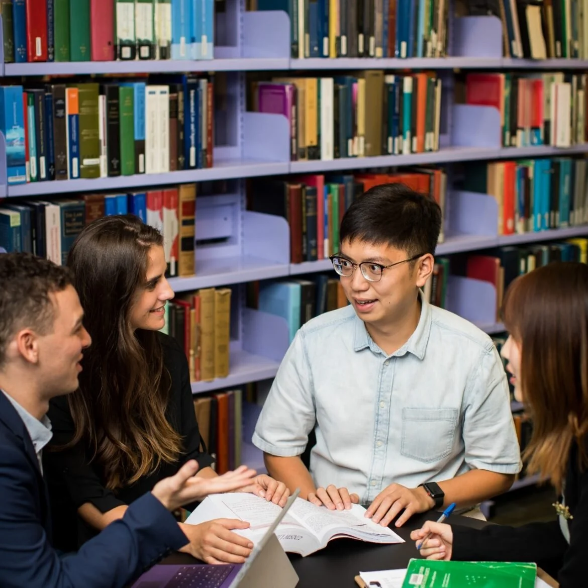 Group of diverse young adults having a discussion in a library with bookshelves in the background.