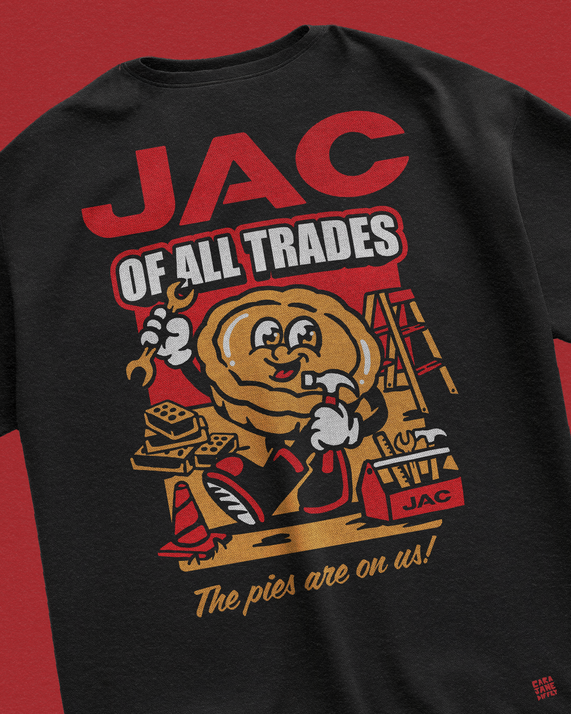 Black t-shirt with a cartoon graphic featuring a pie character holding a wrench, surrounded by tools, pies, a ladder, and construction cones. The text on the shirt reads 'JAG of All Trades' and 'The pies are on us!'