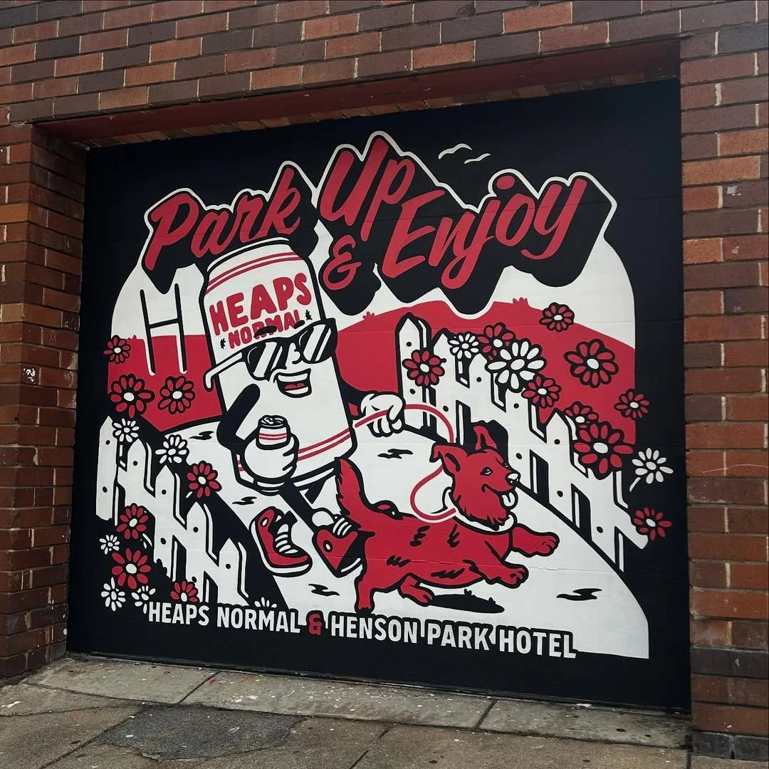 A colorful mural with a cartoon soda can character wearing sunglasses, holding a small container, and walking a dog on a leash along a path lined with flowers. The text reads "Park Up & Enjoy" at the top and "Heaps Normal & Henson Park Hotel" at the 