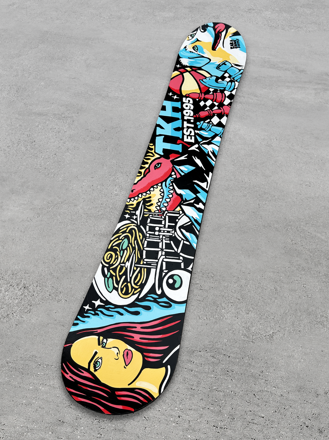 Colorful skateboard deck with graffiti-style artwork featuring a woman with red hair, a dinosaur, a bicycle, a tentacle, and the text 'TXHA EST. 1995'.