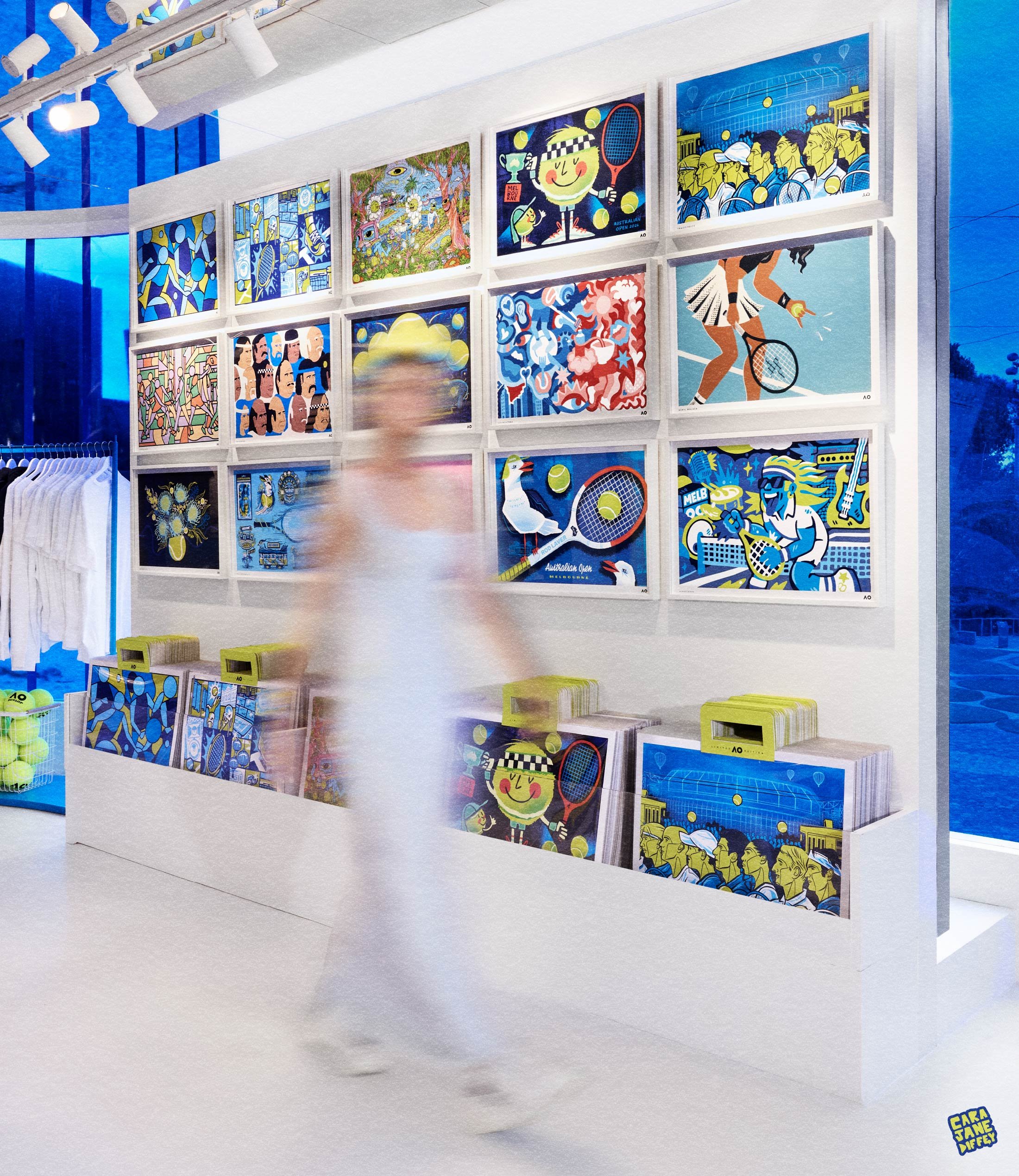 Gallery display of vibrant, colorful artworks with a person walking past, blurred due to motion, in an indoor space with white walls and large windows.