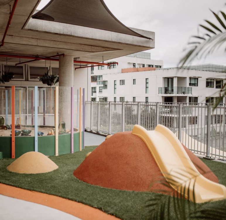 Outdoor play area with colorful hills and a yellow slide on a grassy surface, surrounded by a metal fence, with modern apartment buildings in the background and a canopy overhead.