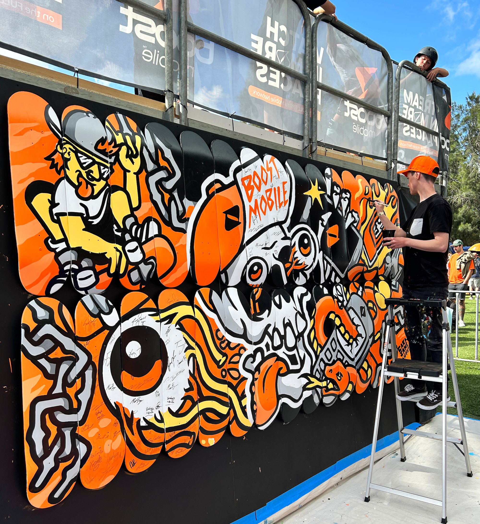 A graffiti mural featuring a cartoonish character wearing a helmet and sunglasses, with bold orange, black, yellow, and white colors. The mural includes a speech bubble that says 'BOOST MOBILE' and a stylized eye with a fiery design around it, with m
