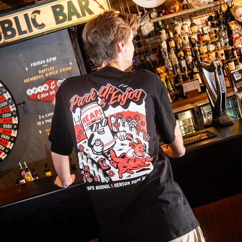 A man stands at a bar counter with a shirt that says 'Park & Enjoy', featuring a cartoon of a dog, a mountain, and flowers. The bar has various bottles of alcohol and a Guinness beer tap. There's a blackboard behind the man with a dartboard illustrat