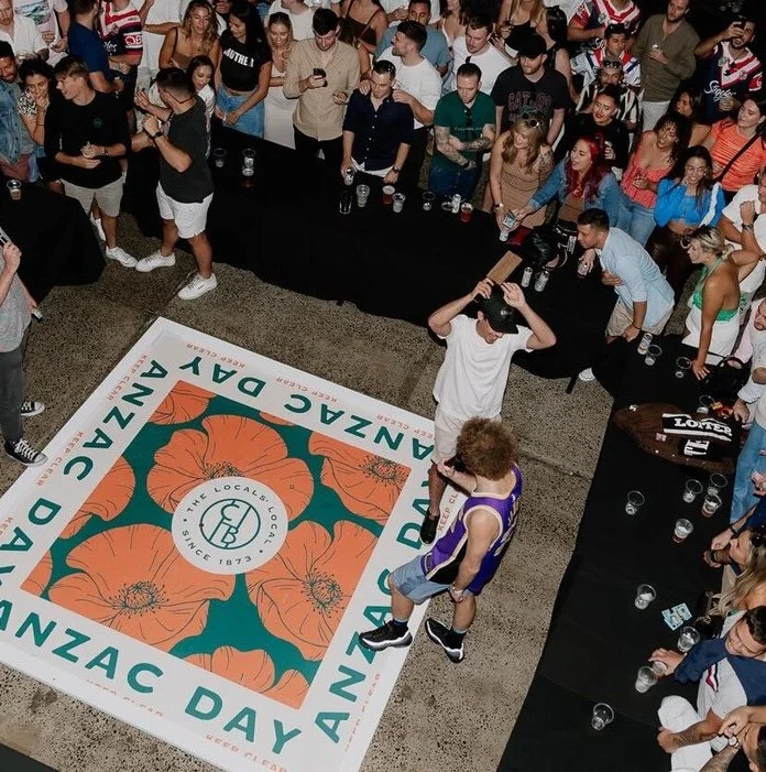 A large group of people gathered around a table and a decorative banner on the floor, with one person in a purple shirt and shorts holding an award or plaque, at an event celebrating Anza Day.