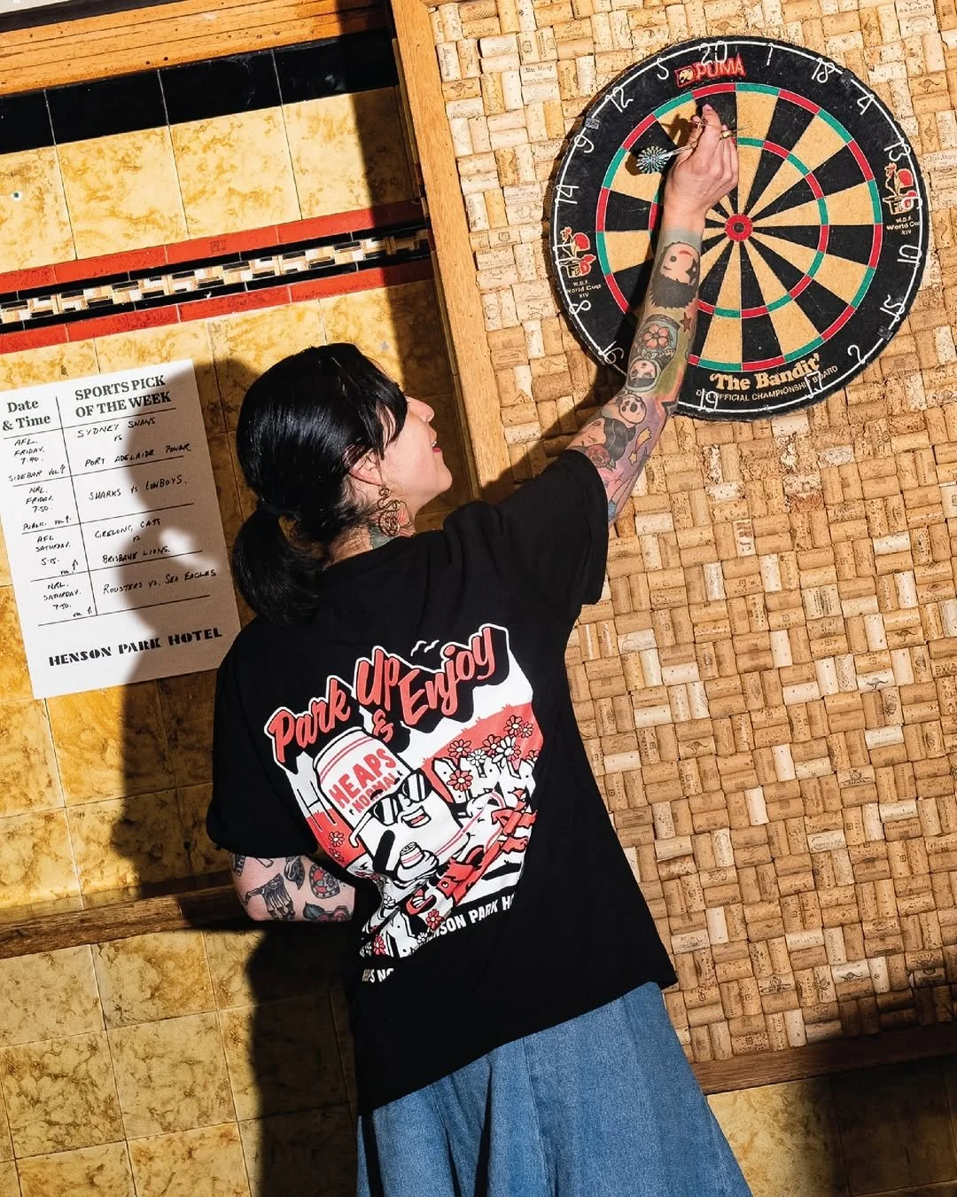 Person with tattooed arm wearing a black T-shirt and blue jeans playing darts in a bar or pub setting.