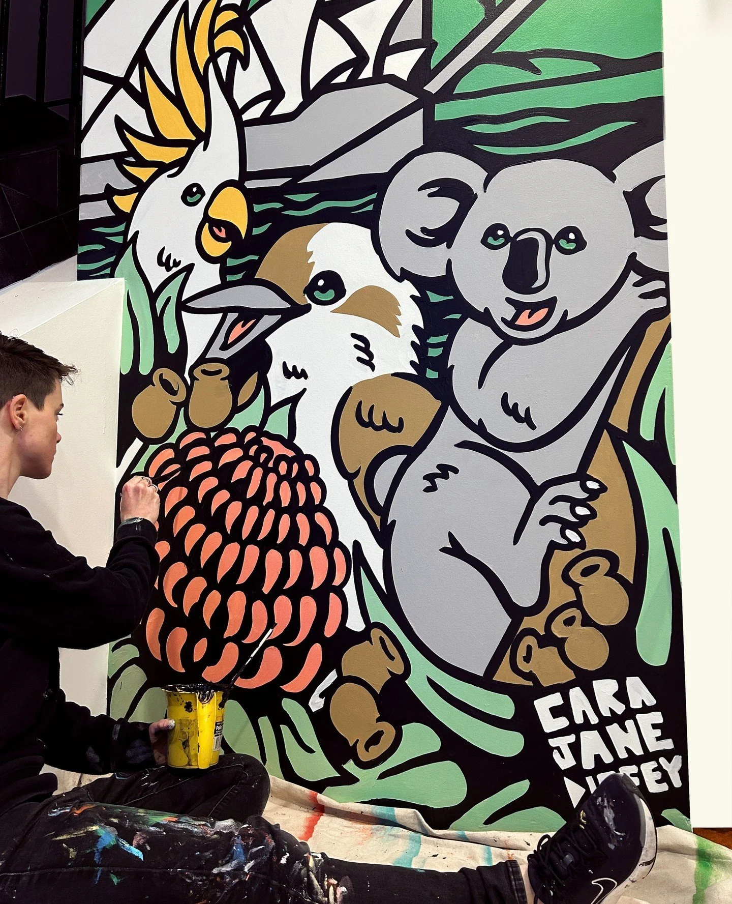 A person painting a colorful, cartoon-style mural of a cockatoo, a duck, and a koala, with the artist sitting on the floor holding yellow paint cans and working on the artwork.