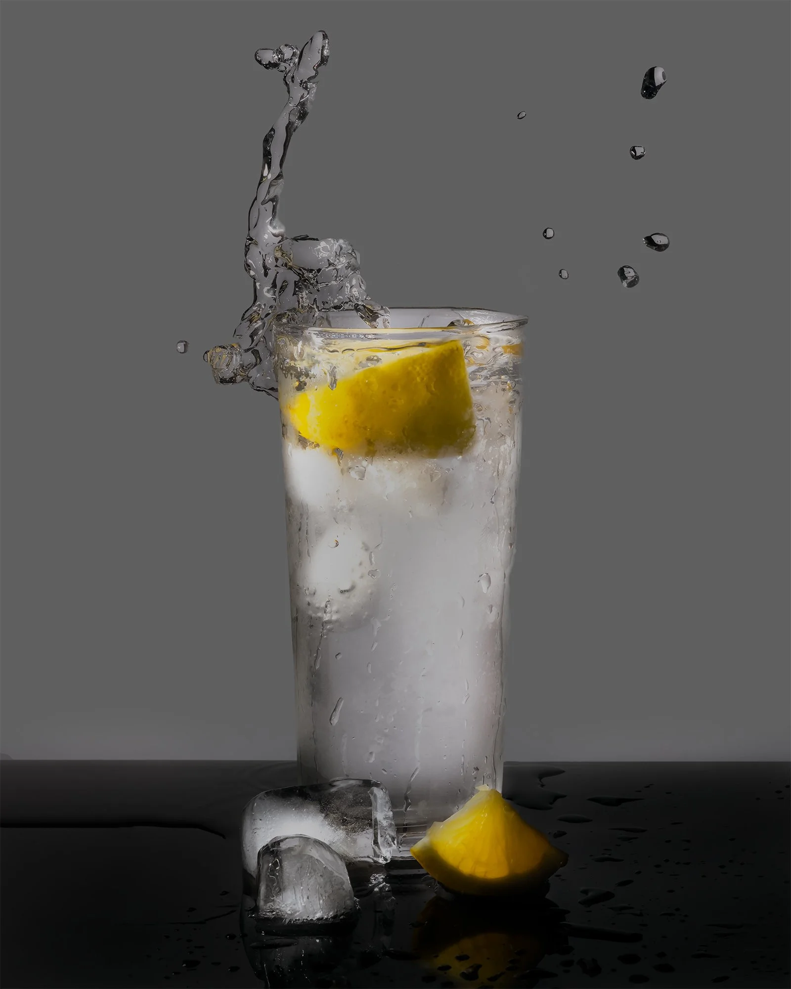 A tall glass of water with lemon slice, ice cubes, and splashing water against a gray background.