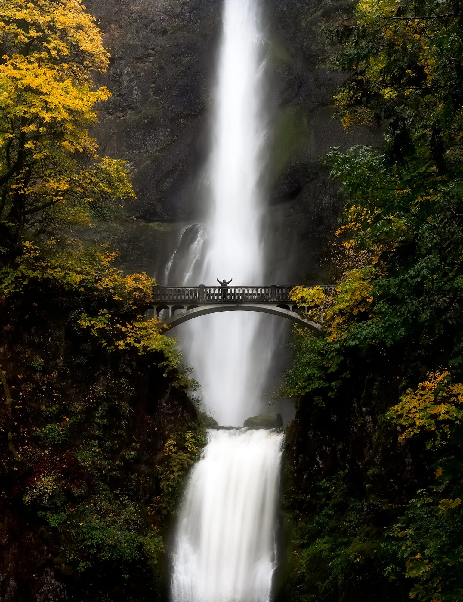 A two-tiered waterfall with a person standing on a footbridge in front of it, surrounded by autumn-colored trees.