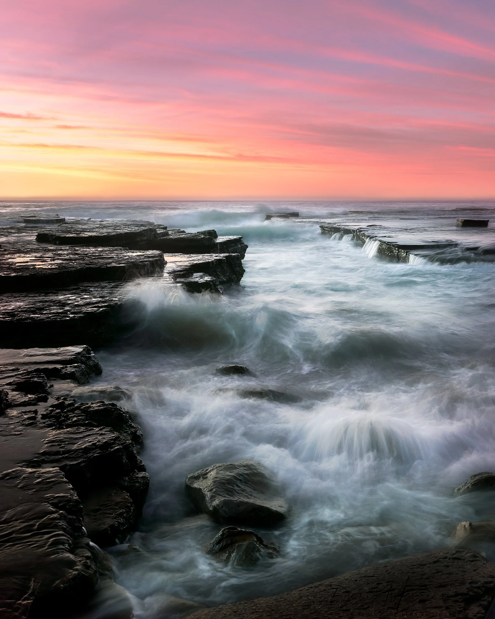 Twilight over rocky ocean shore with waves crashing against rocks and a colorful pink and orange sunset sky.