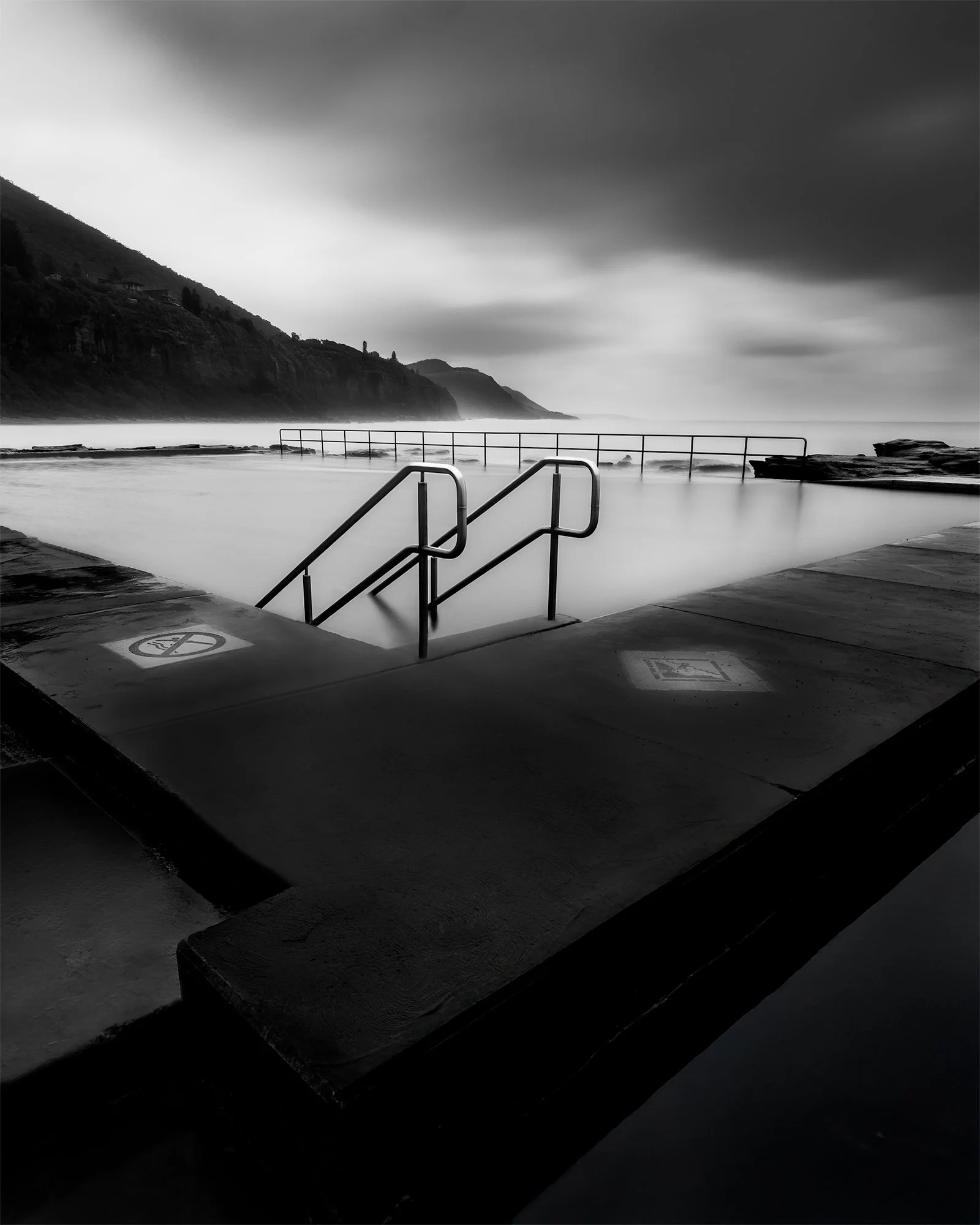 Black and white photograph of an empty outdoor swimming pool with metal handrails and warning signs on the edge, overlooking a foggy ocean with hills in the background.