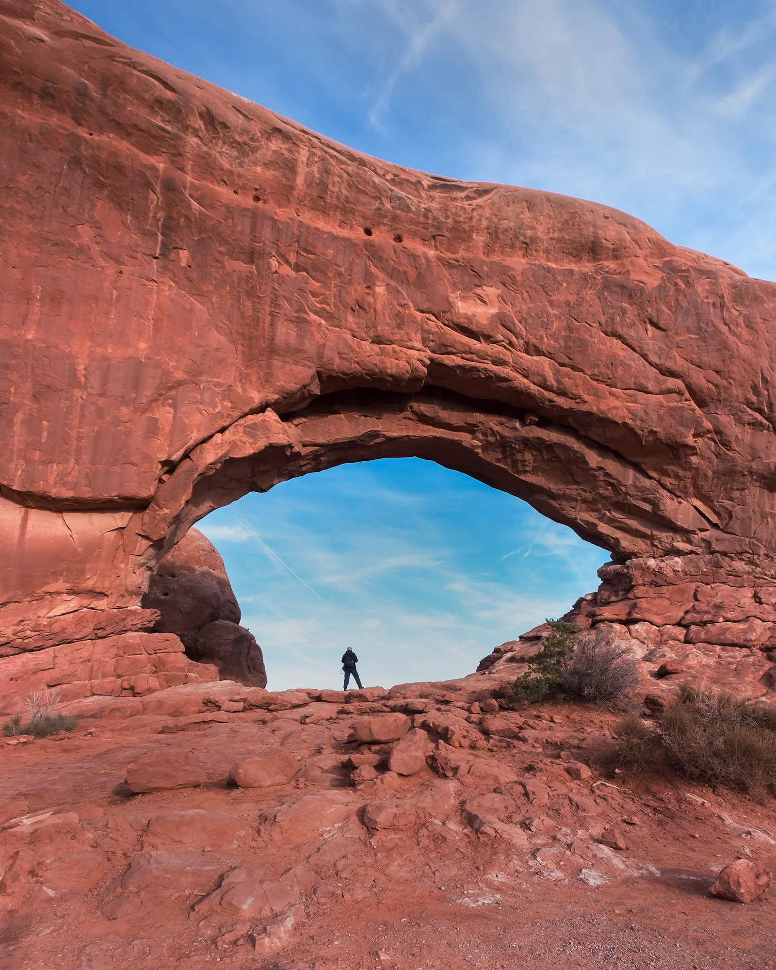 A person standing under a large natural red rock arch in a desert landscape with a blue sky.