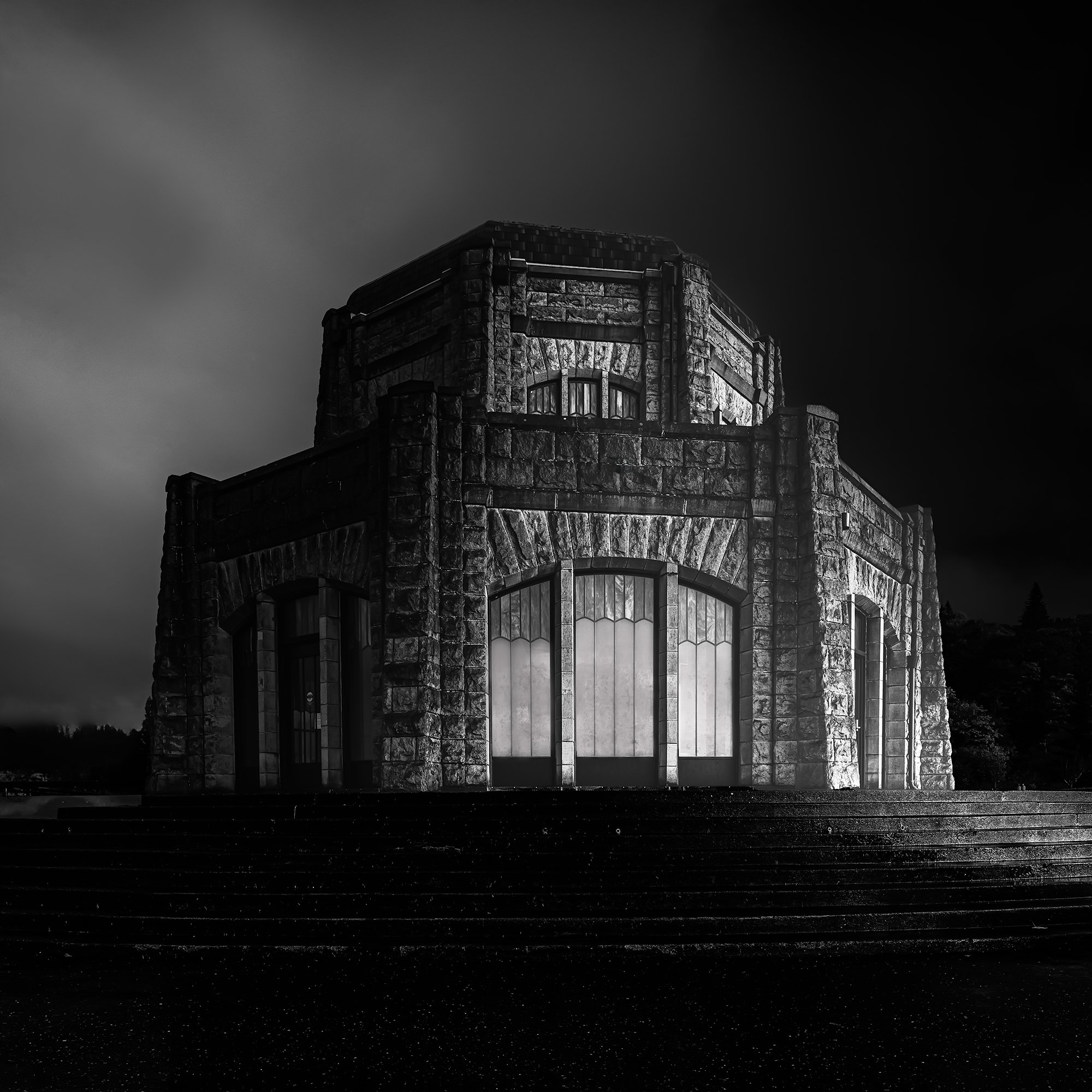 A large stone building with gothic architecture, glowing lights visible inside through large arched windows, under dark sky.