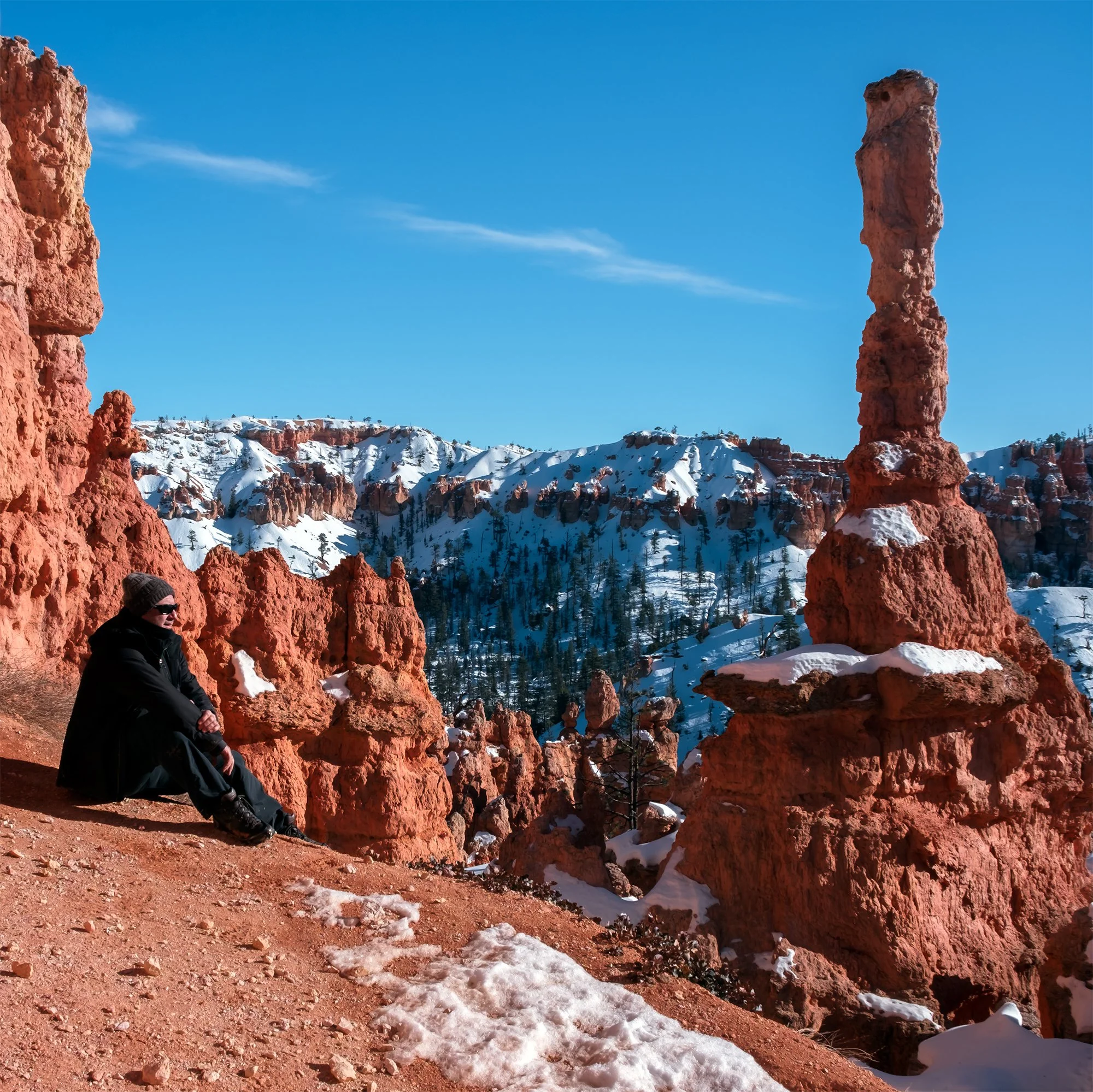 A person wearing winter clothing sitting on a rocky ledge in a snowy canyon with hoodoos and snow-covered mountains in the background on a clear, sunny day.