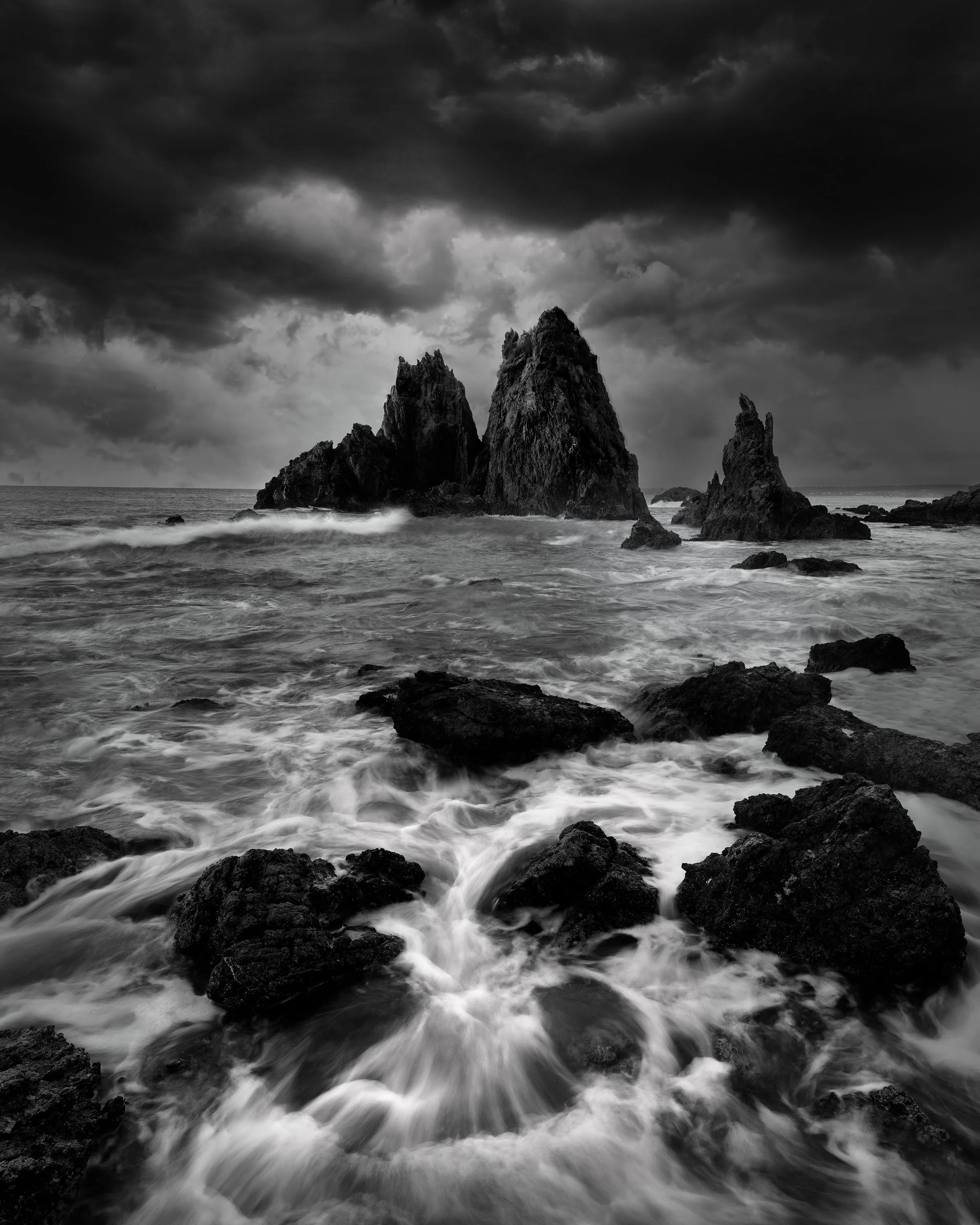 Black and white photograph of rocky sea stacks and turbulent ocean waves under a dramatic cloudy sky.