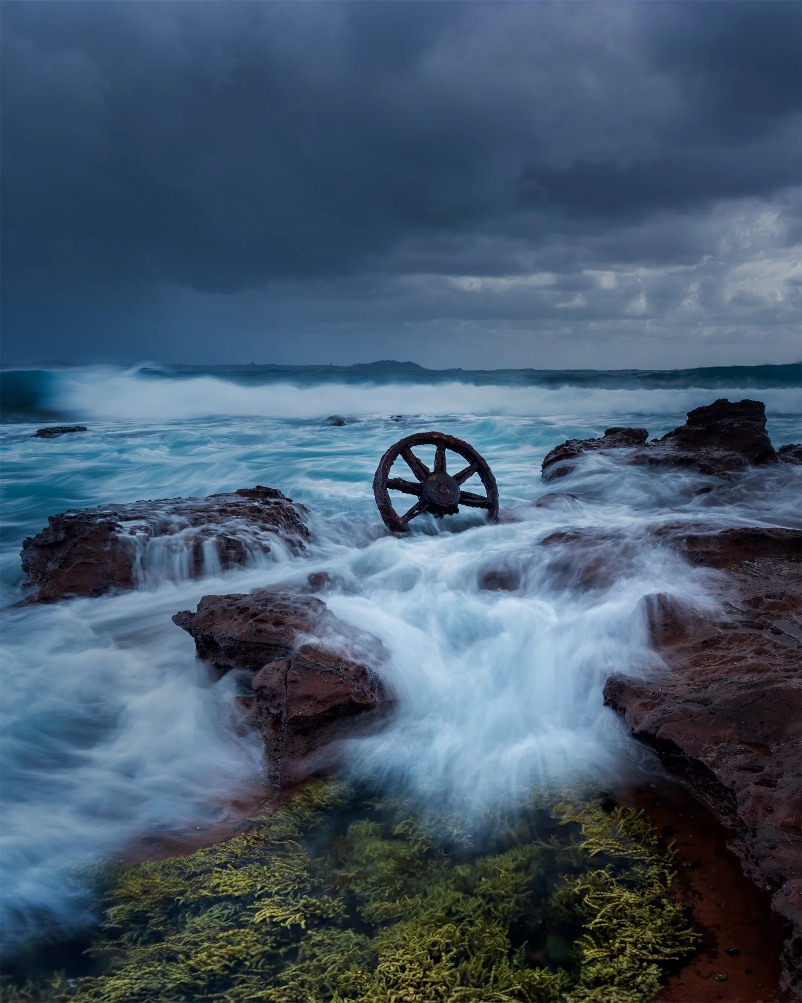 Sea waves crashing over rocks with an old rusted ship wheel submerged in water under a dark cloudy sky.