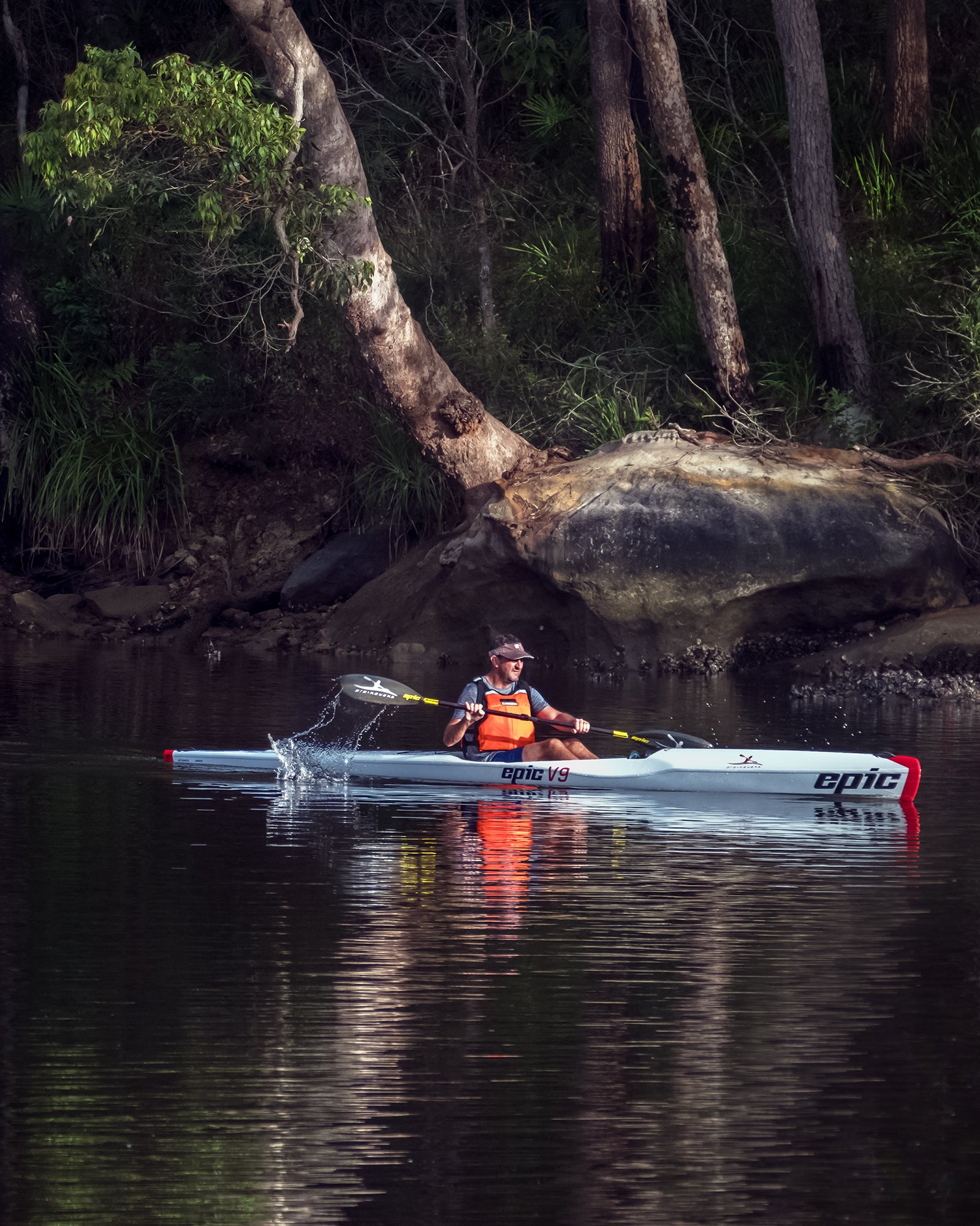 A man kayaking on a calm body of water near a forested shoreline with large rocks and trees.