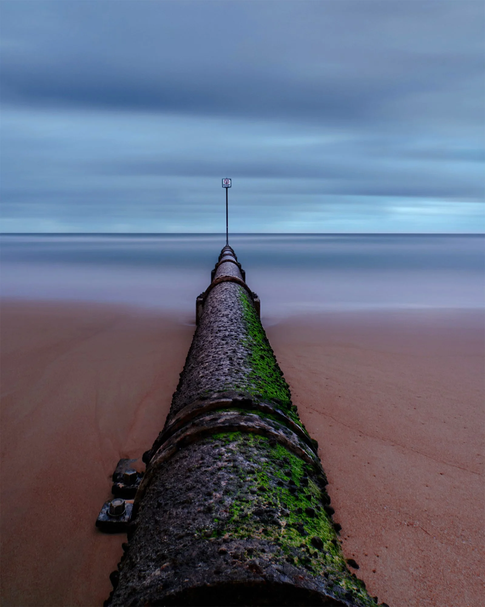 An old, rusted pipe with green moss running along a sandy beach, extending into the ocean with a sign at the end of the pipe, under a cloudy sky.