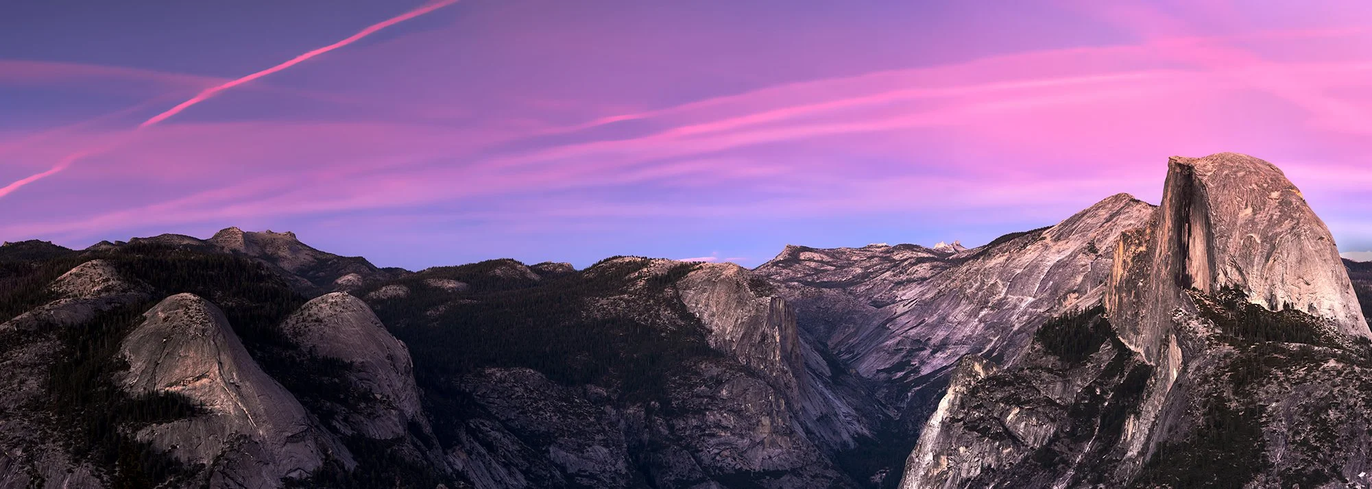 Pink and purple sunset over a mountain landscape with large granite rock formations and forested areas.