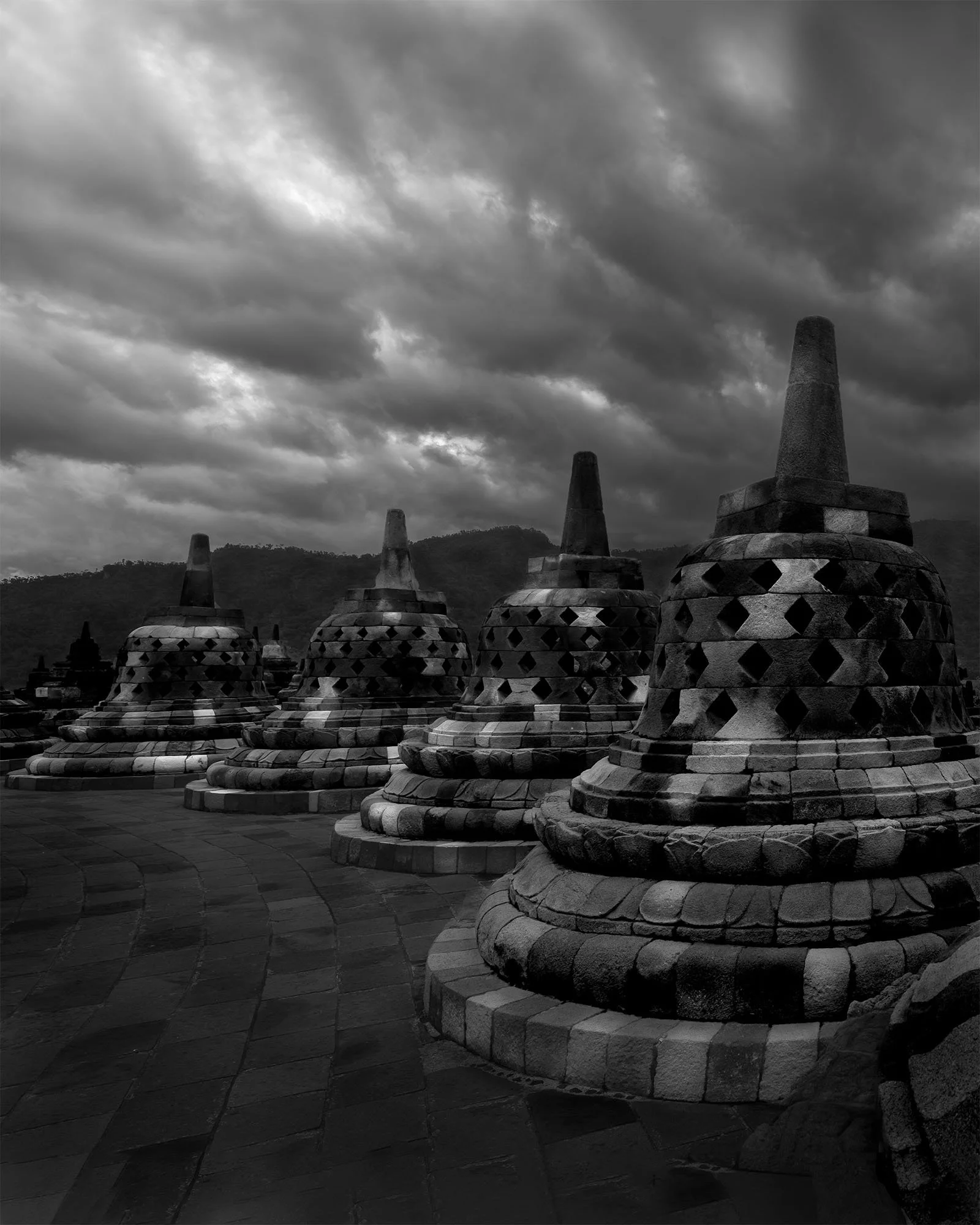 Four large stone stupas with patterned exteriors on a paved platform, set against a cloudy sky and mountainous backdrop.