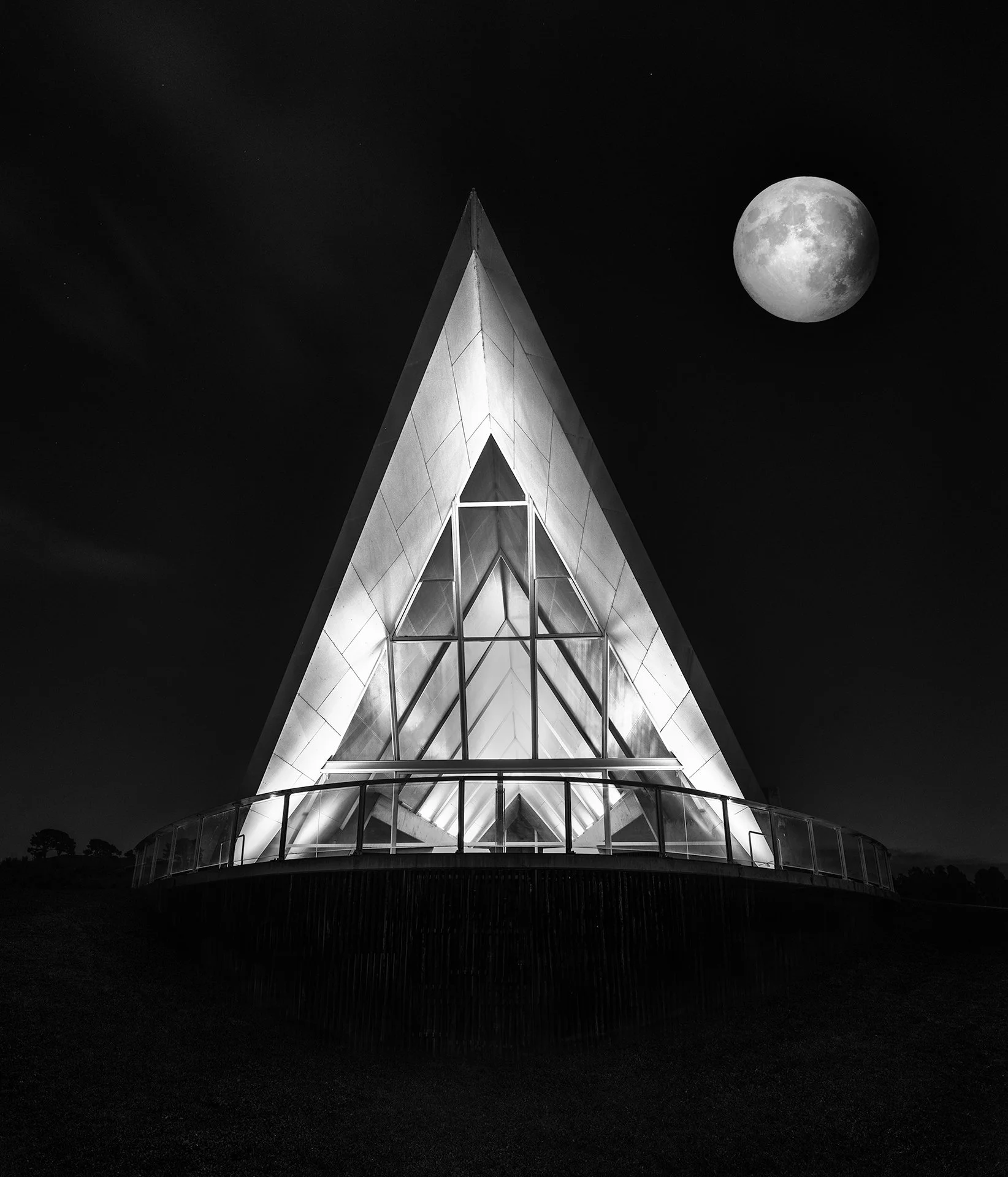 A modern triangular glass building illuminated at night with the full moon in the sky above.