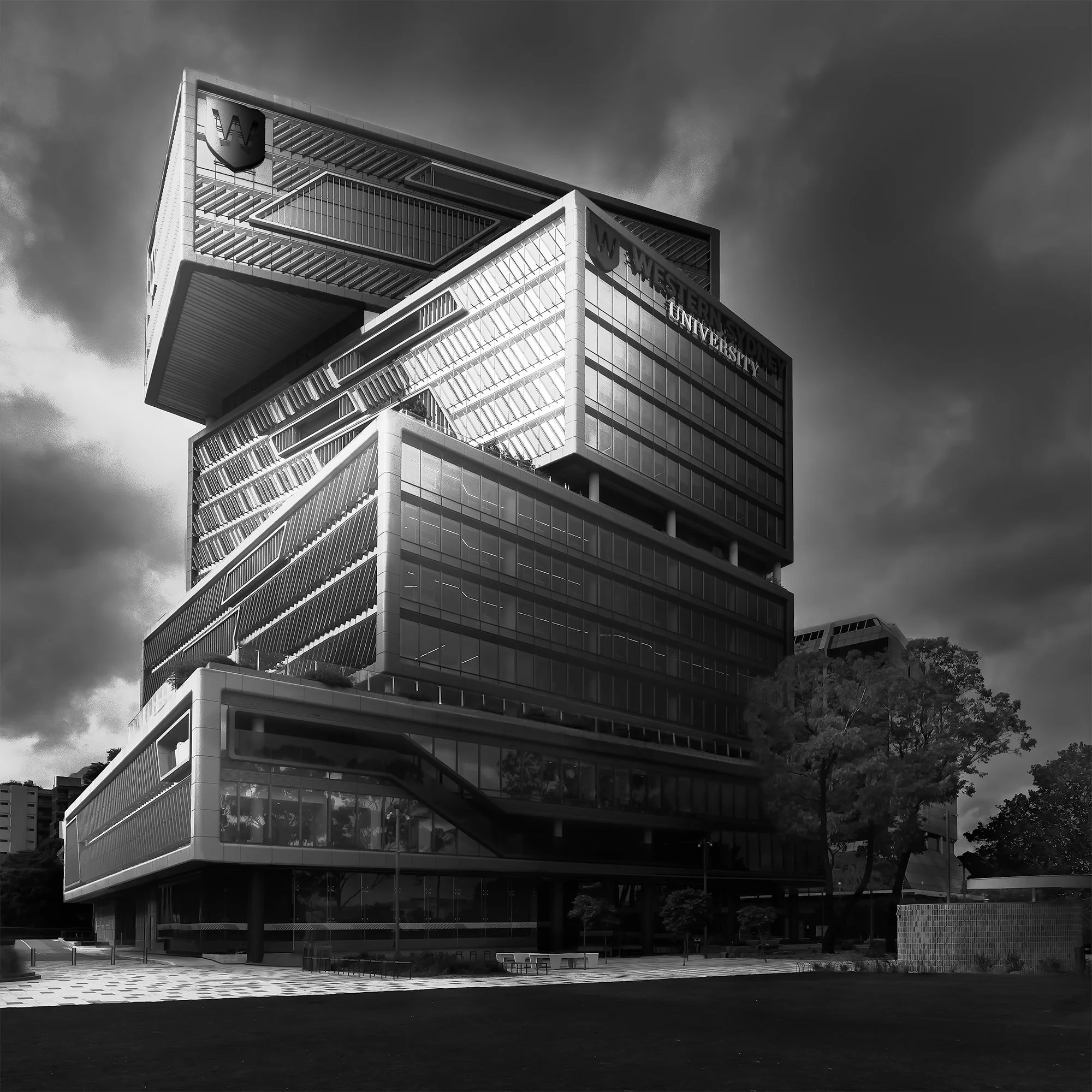 A black-and-white photograph of a modern multi-story university building with the sign 'Western Sydney University' at the top, featuring a distinctive architectural design with glass and steel elements, trees, and an open paved area in front.