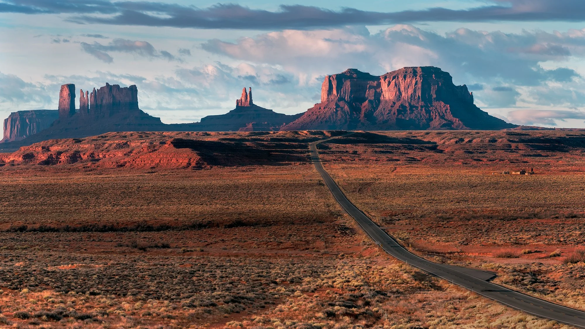 A long road stretches through a desert landscape leading to rock formations and buttes in Monument Valley, with a partly cloudy sky overhead.