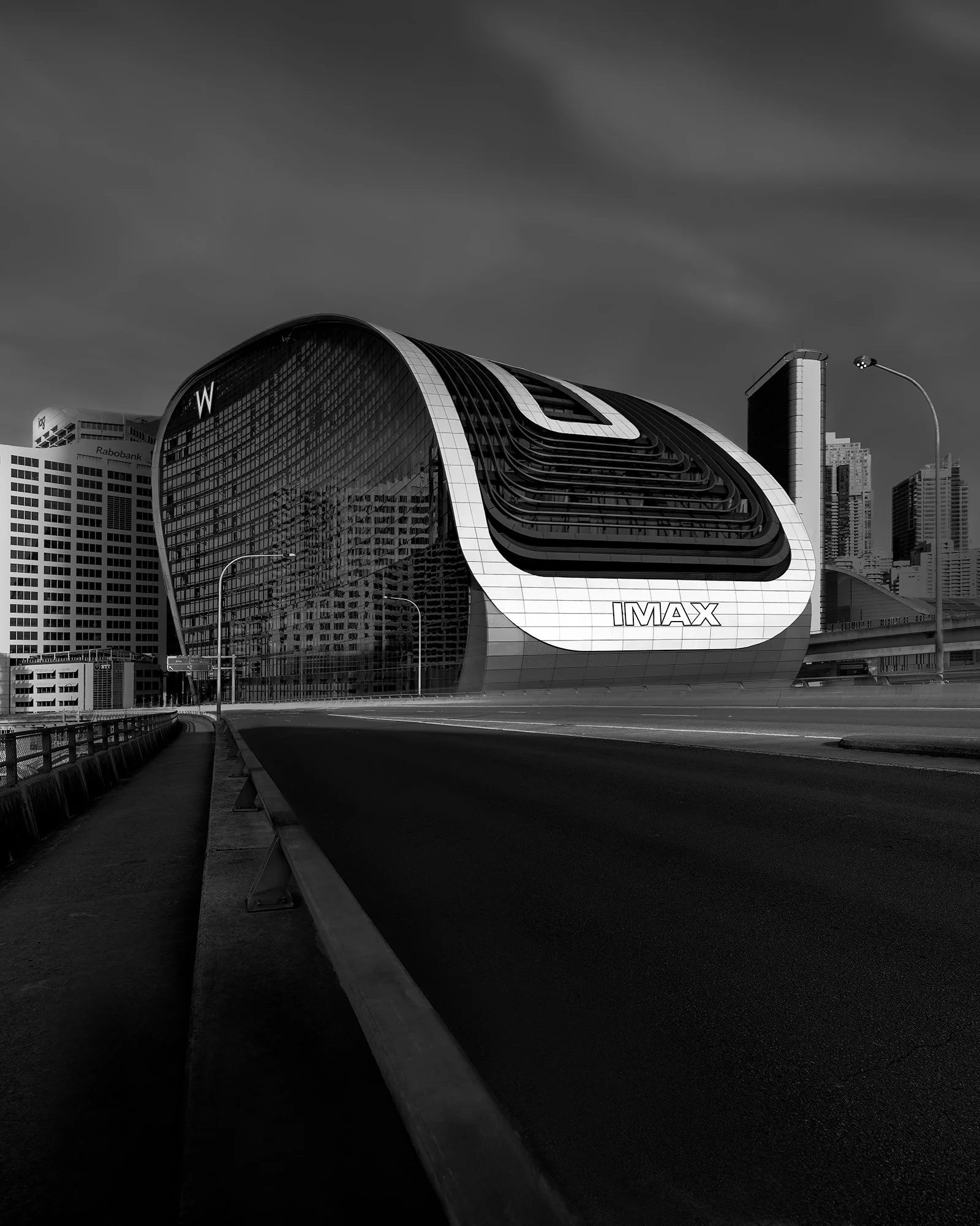 Modern futuristic building in an urban cityscape with the IMAX logo on its front, under a cloudy sky in black and white.